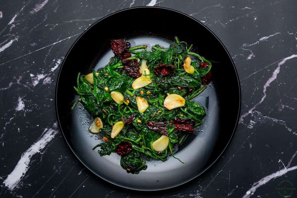 A black bowl filled with Wilted Spinach, Garlic Slices, and Chilis